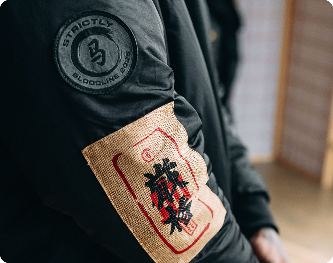 Person wearing a black jacket with a brand logo on the sleeve and a burlap patch with red and black text on the arm.